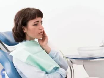 This shows a patient with clear signs of pain on the left side of her mouth in a dentist chair {{brizy_dc_image_alt entityId=